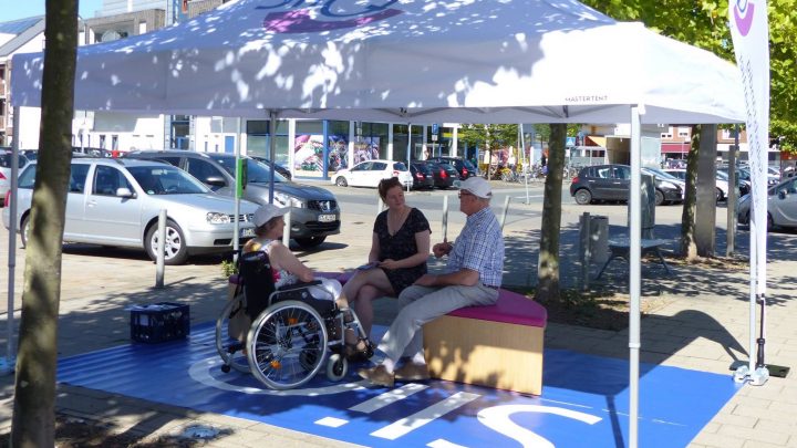 Auf dem Foto sitzen drei Menschen, einer davon im Rollstuhl, unter einem Pavillon in einer Fußgängerzone und sprechen miteinander.